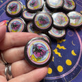 Collection of buttons with unicorn designs saying Bad Bitch held by a hand on a starry night background