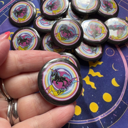 Collection of buttons with unicorn designs saying Bad Bitch held by a hand on a starry night background